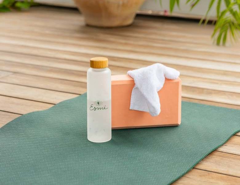 a white bottle with a wooden cap and the word 'Esmé' on it, a peach-colored box with a white towel on top, and a green mat on a wooden surface