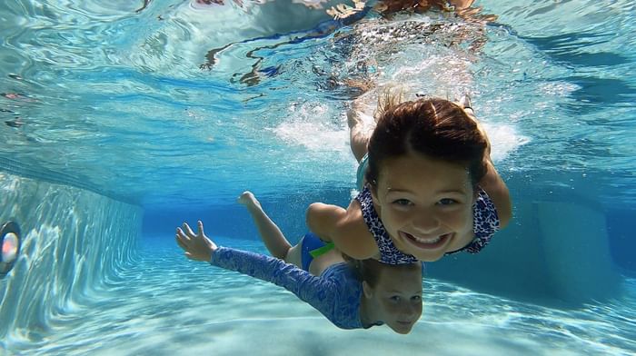 Underwater view of two happy young girls smiling while swimming in the bright, clear resort pool at Centennial Plaza Resort