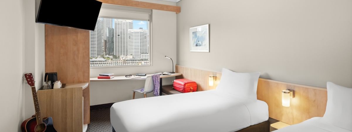 Superior room with two single beds, work desk, wall-mounted TV, and city skyline view at Ibis Sydney Darling Harbour
