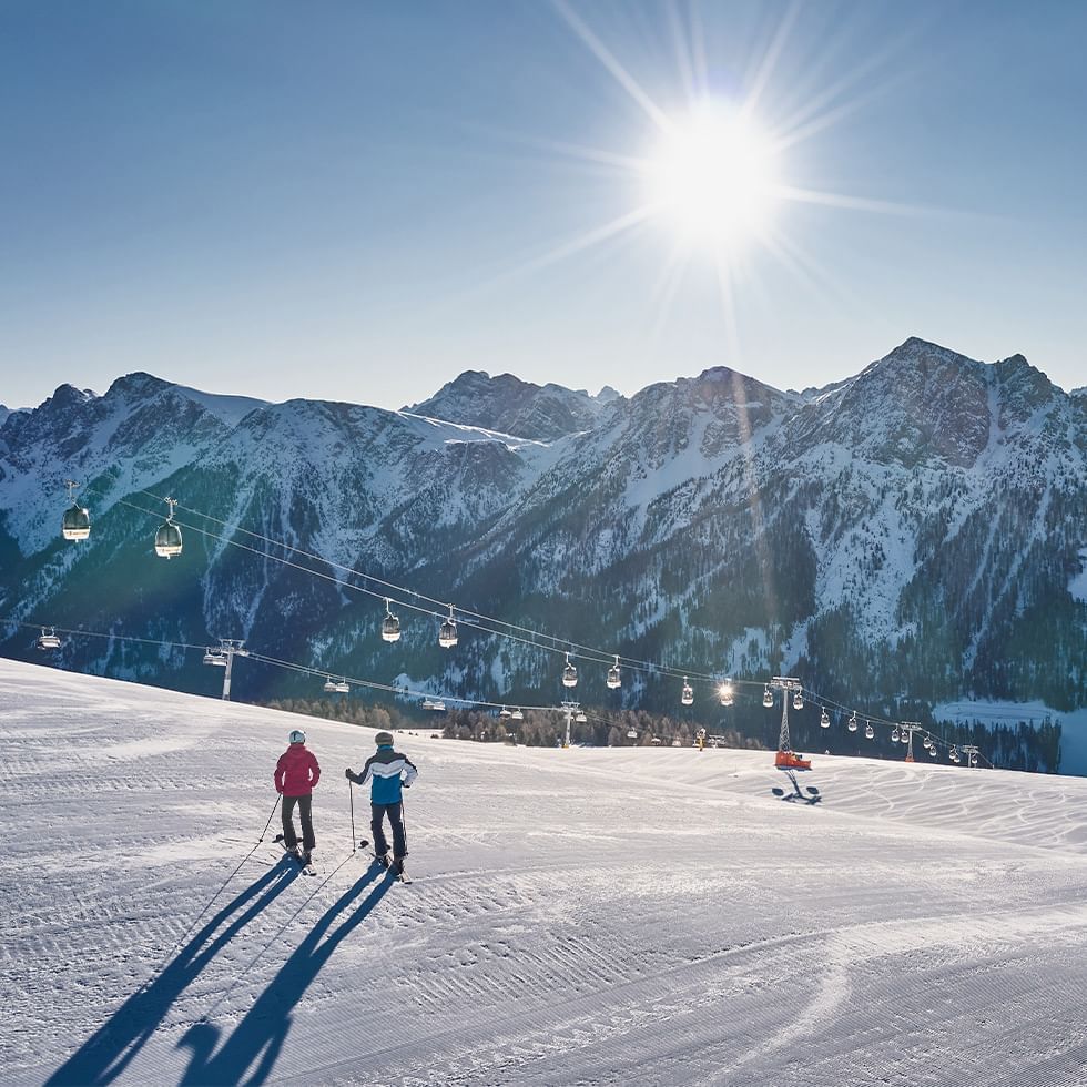 Zwei Skifahrer auf einer schneebedeckten Piste mit Bergpanorama und Gondelbahnen im Hintergrund.