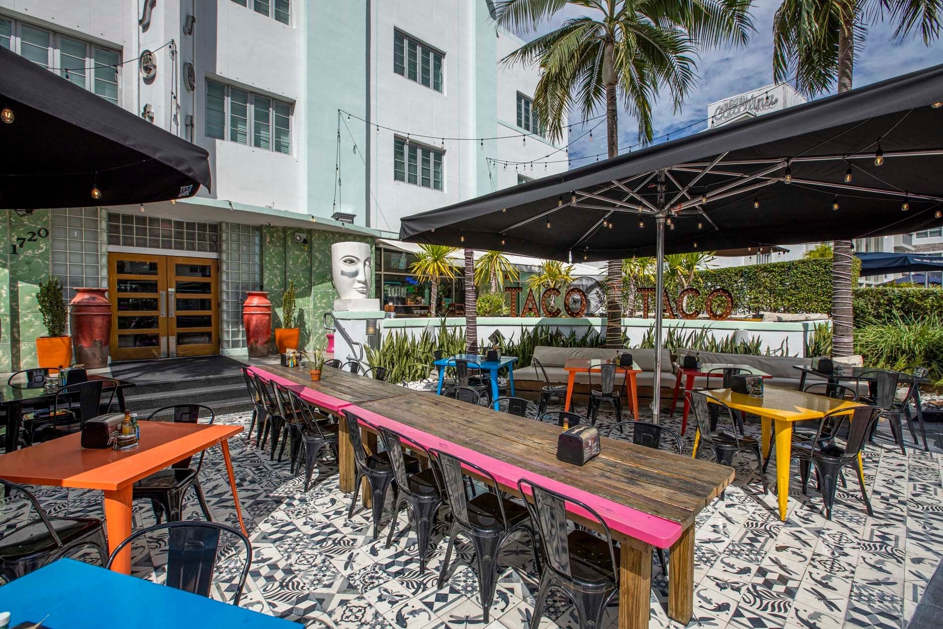 A colorful outdoor dining area with tables and chairs under umbrellas, surrounded by palm trees and a building in the background.