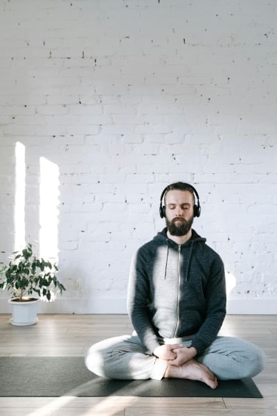 A bearded man in a white room sits on a yoga mat with his legs crossed and his eyes closed. 