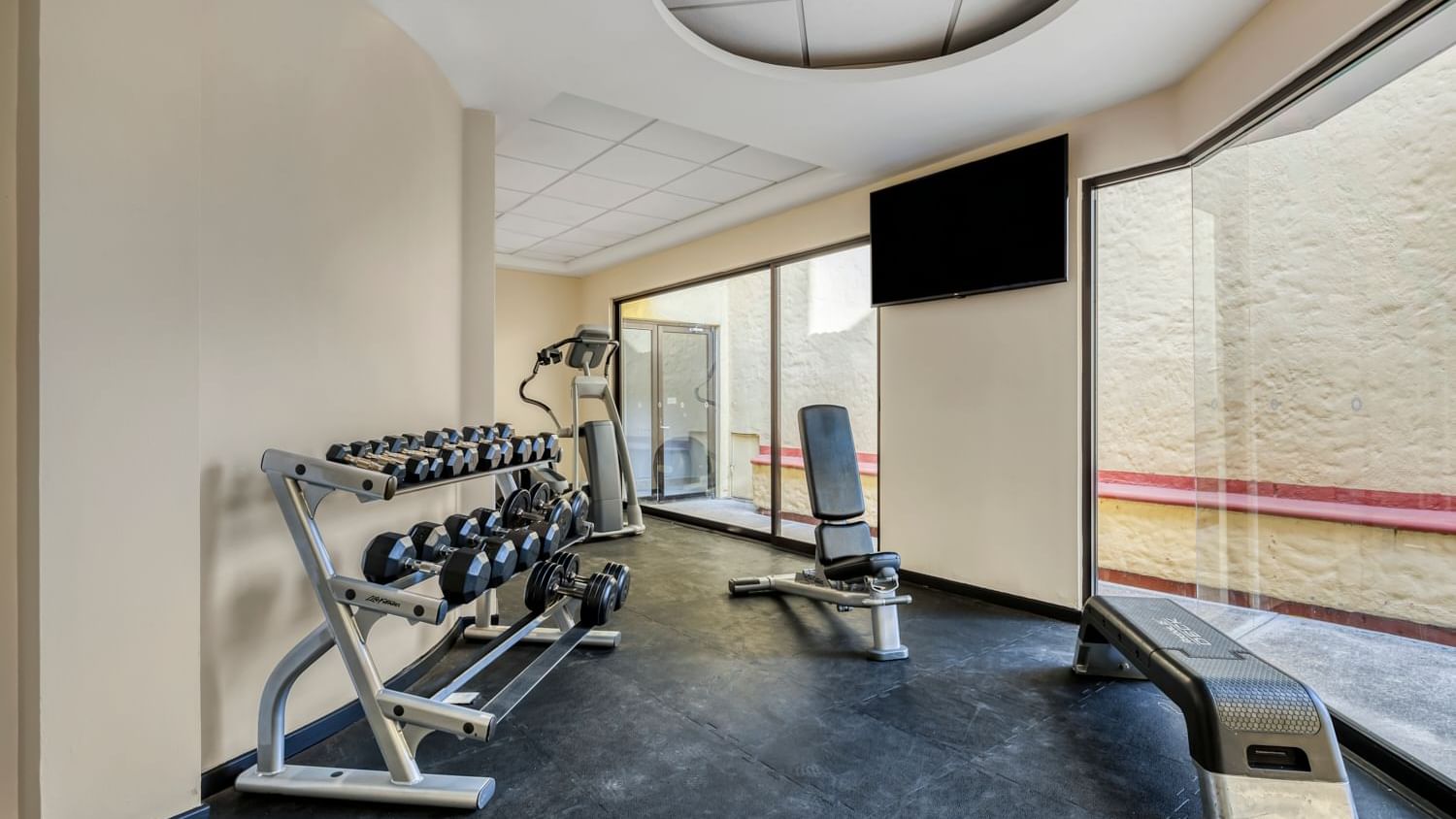 Gimnasio con mancuernas, banco de pesas y televisor en la pared junto a ventanas de vidrio.