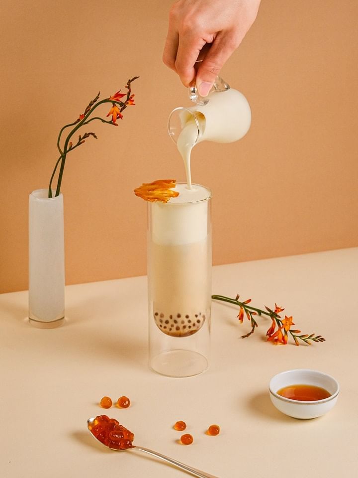 Hand pouring milk into a layered bubble tea with dried fruit, surrounded by orange flowers in Park Lounge, Park Hyatt Saigon