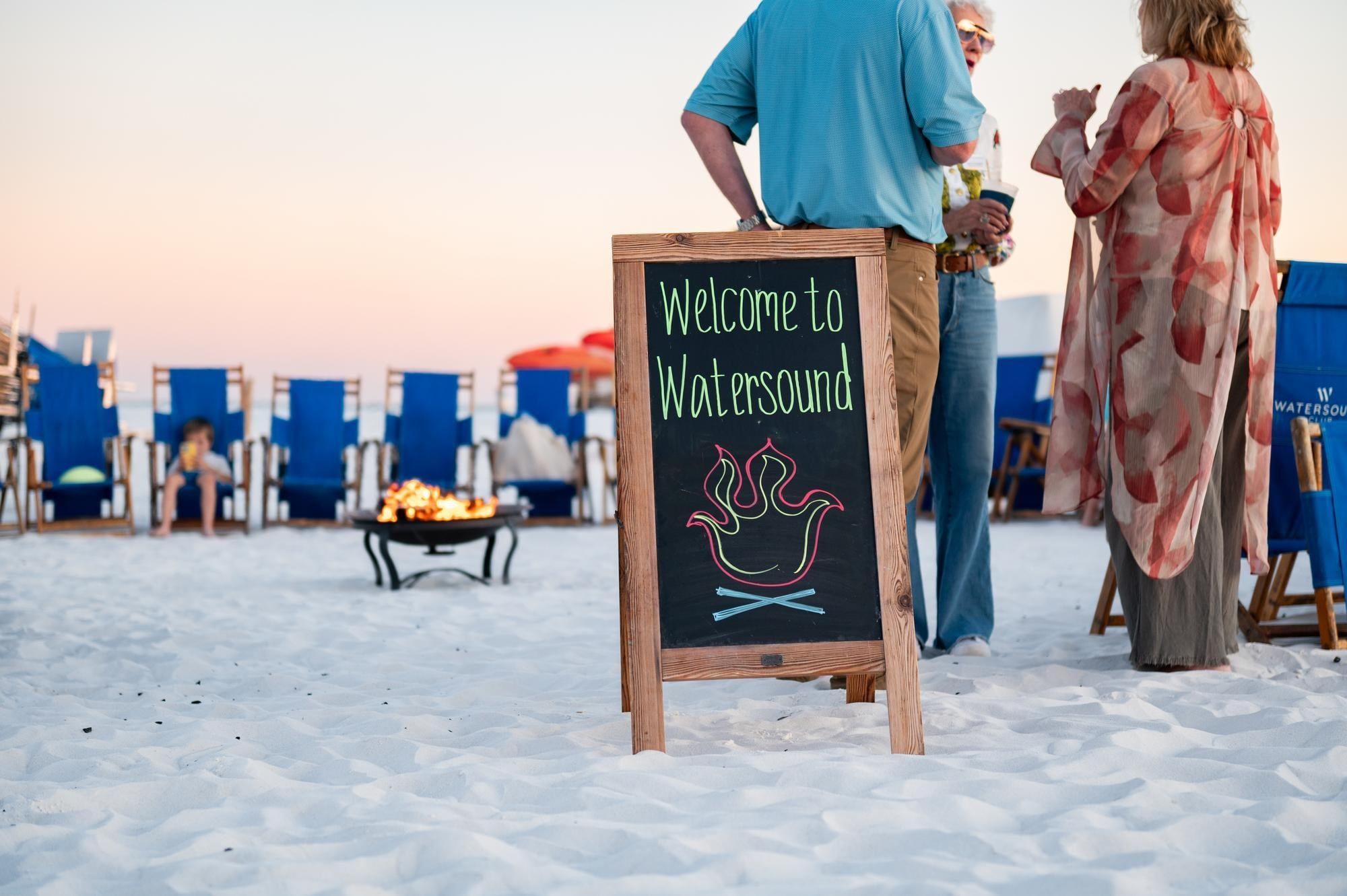 Beach gathering with welcome sign, blue chairs, fire pit, and guests enjoying sunset at Camp Creek Inn