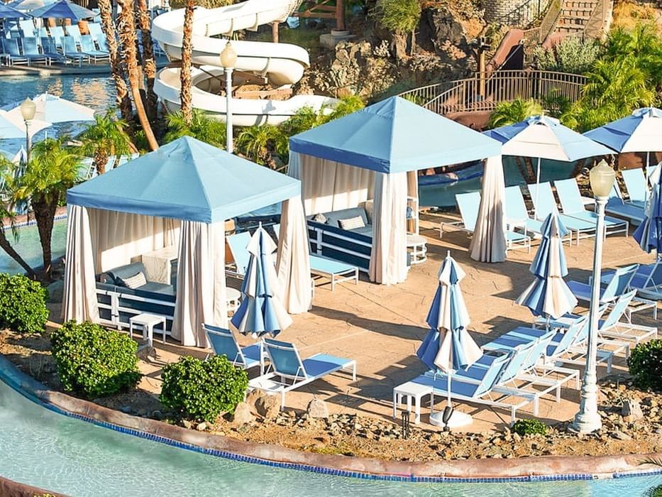 Aerial view of a resort pool deck with blue cabanas, lounge chairs, and a lazy river surrounded by palm trees and greenery