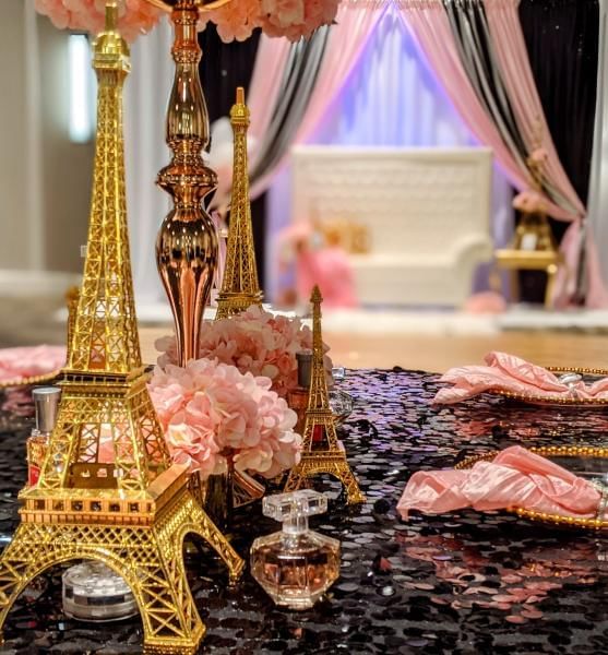 A table setting with gold Eiffel Towers, a black sequined tablecloth, pink linens, and perfume bottles.