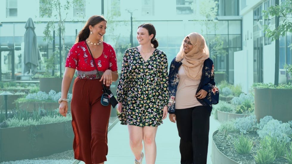 Three women walking together on a pathway, with one wearing a hijab and another in a floral dress.