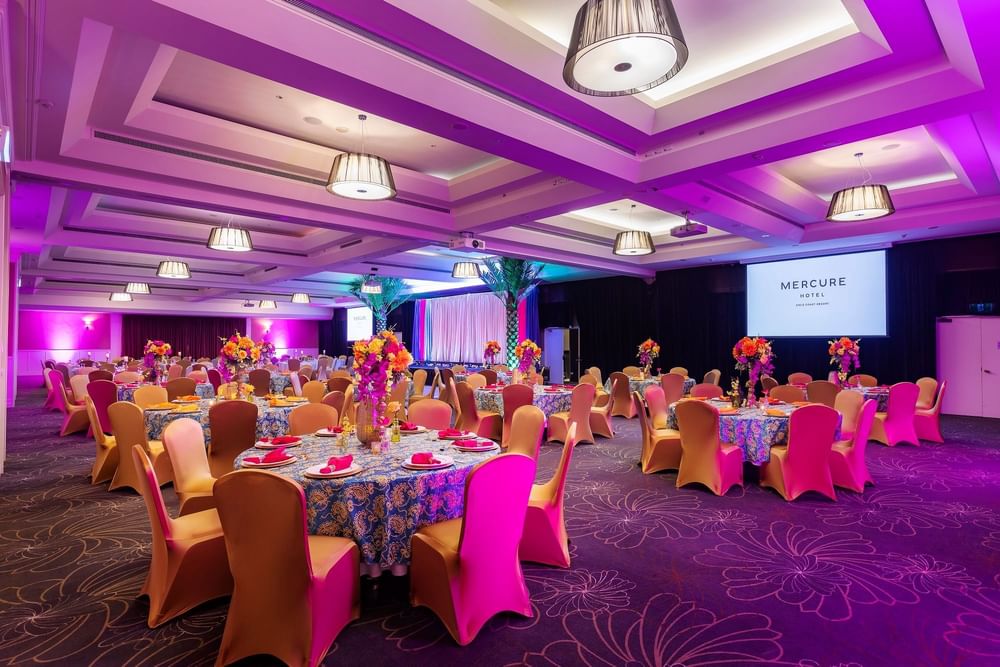 Banquet hall with purple lighting, decorated tables, and chairs at Mercure Hotel, with a large screen displaying hotel name.