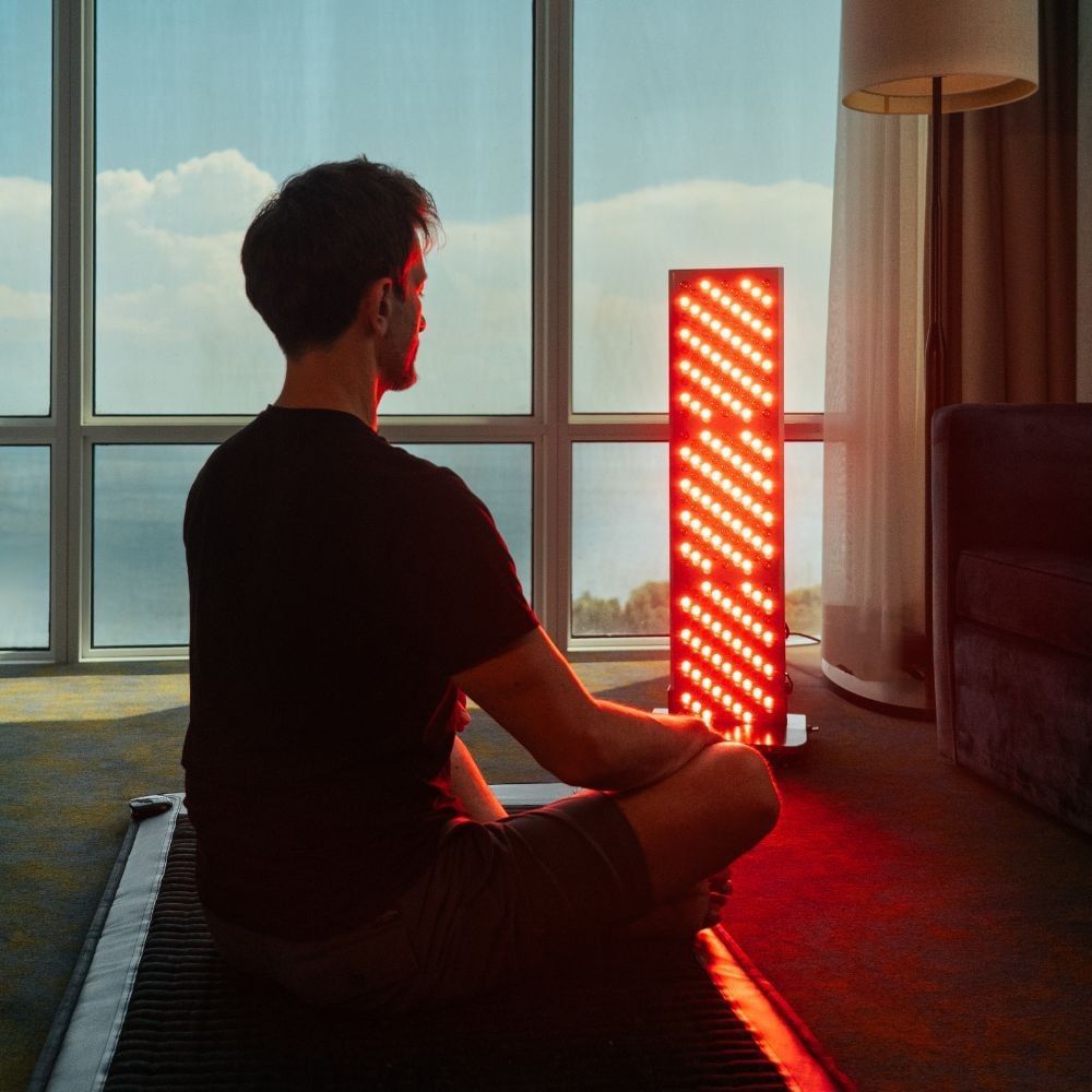 Man meditating beside a glowing red light therapy panel near floor-to-ceiling windows in a guest room at Hotel X Toronto