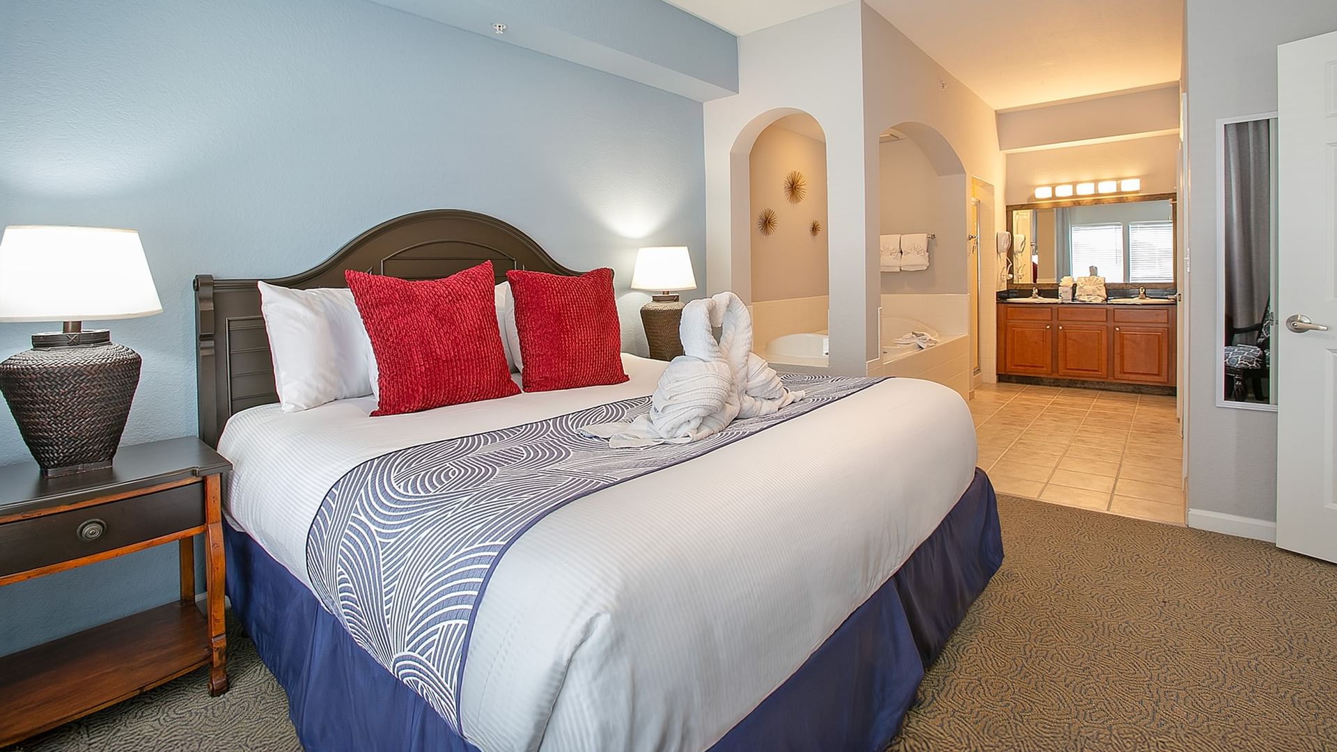 A hotel room with a king-sized bed, nightstand, and lamp, with a tiled bathroom in the background.