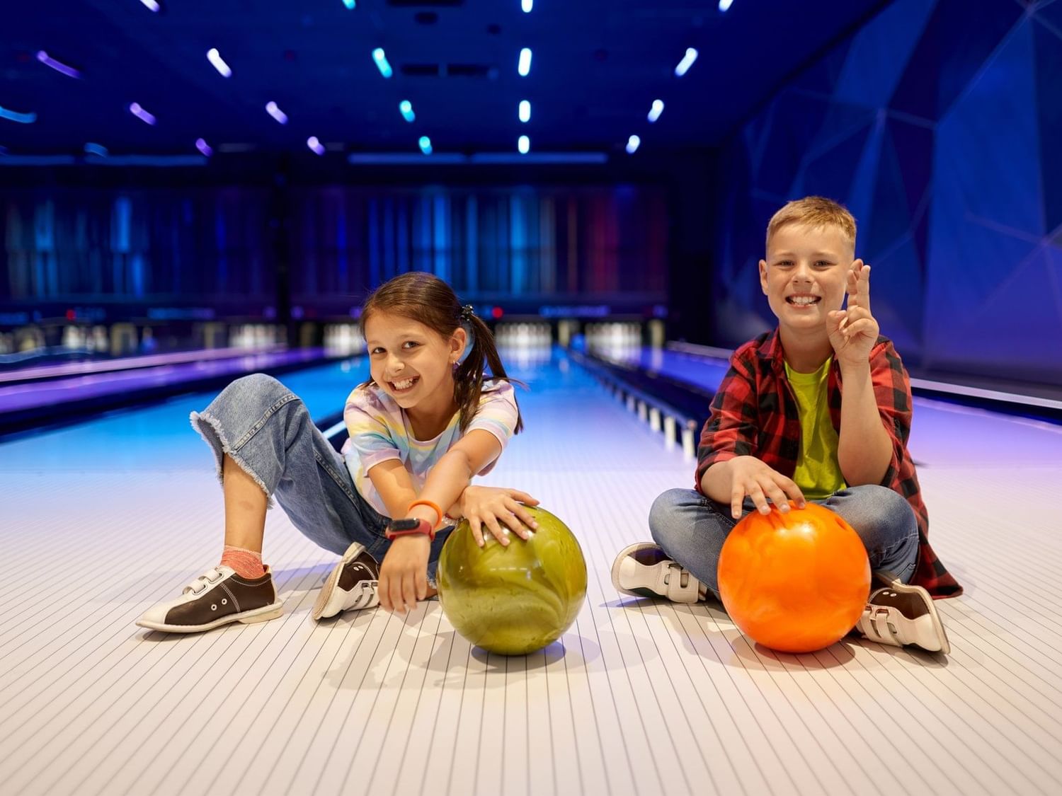 Dos niños felices sentados en una pista de bolos con bolas coloridas en la divertida bolera de Fiesta Americana Funeeq