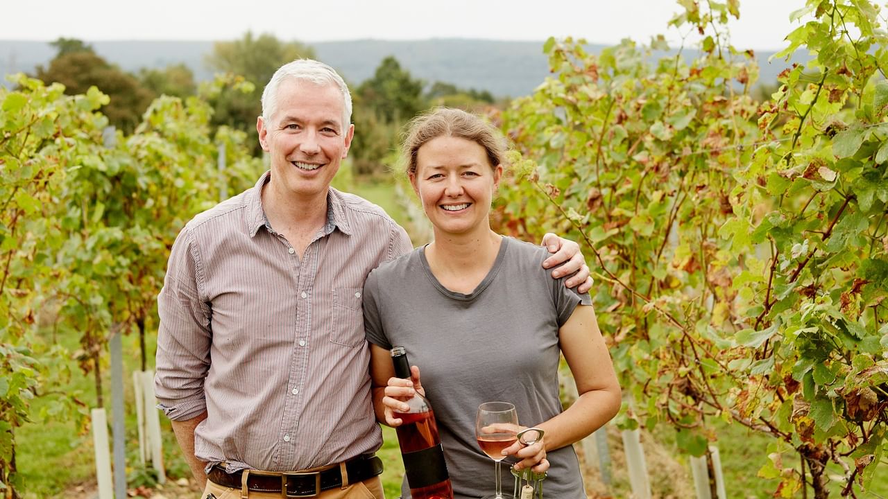 a smiling couple holding wine bottle and glass standing in vineyard