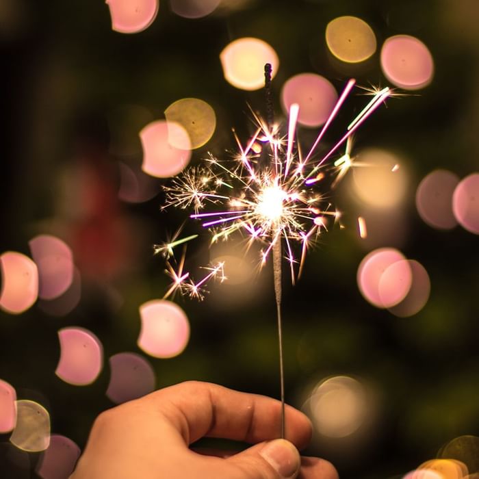 Mano che tiene un petardo acceso con scintille e sfondo sfocato di luci colorate.