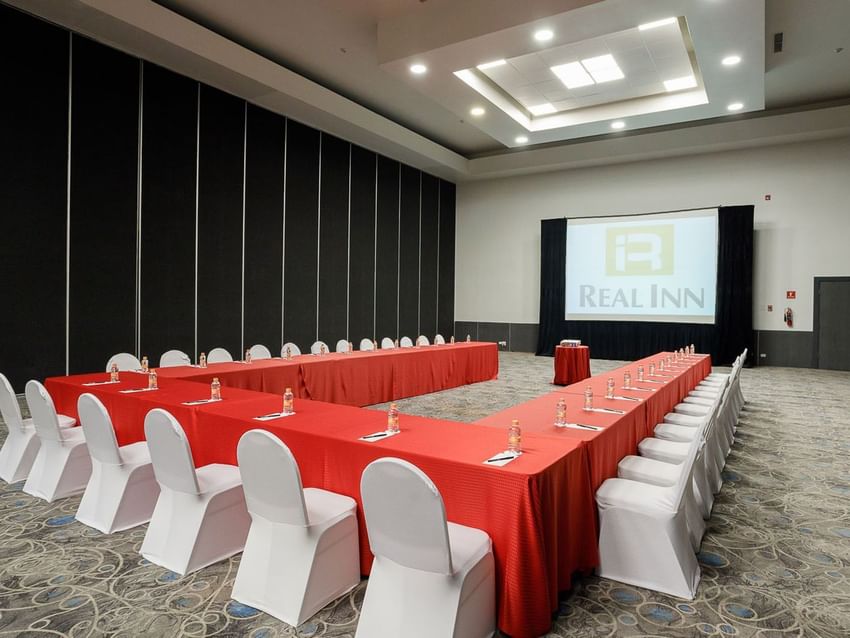 U-shaped meeting setup with red tablecloths and a large projector screen in Venecia Venue at Real Inn Tijuana
