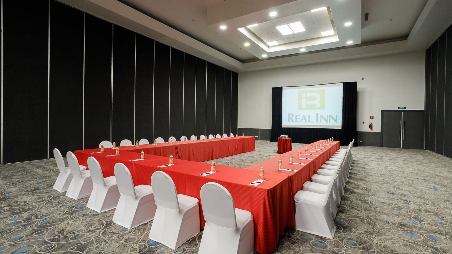U-shaped meeting setup with red tablecloths and a large projector screen in Venecia Venue at Real Inn Tijuana