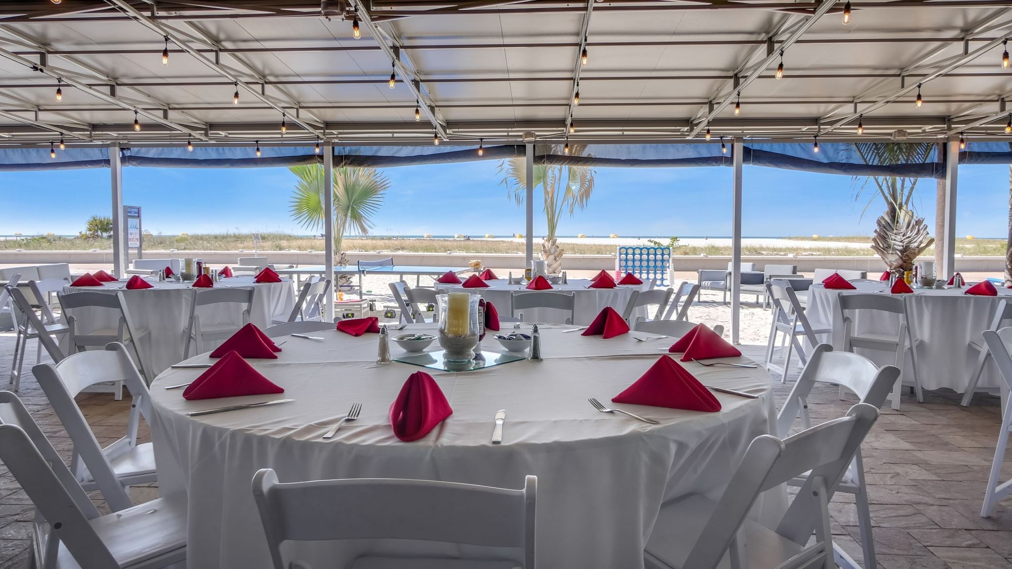 a dining area with white tables and red napkins under a covered patio with a beach view