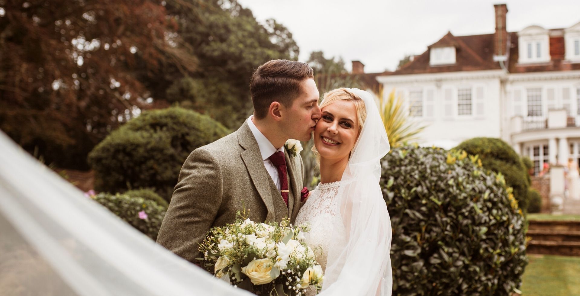 Groom kissing his bride in front of mansion house at Gorse Hill