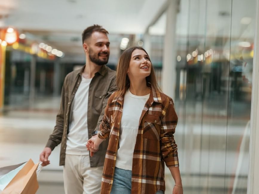 Couple shopping in mall, holding hands and carrying bags, passing by a glass storefront.