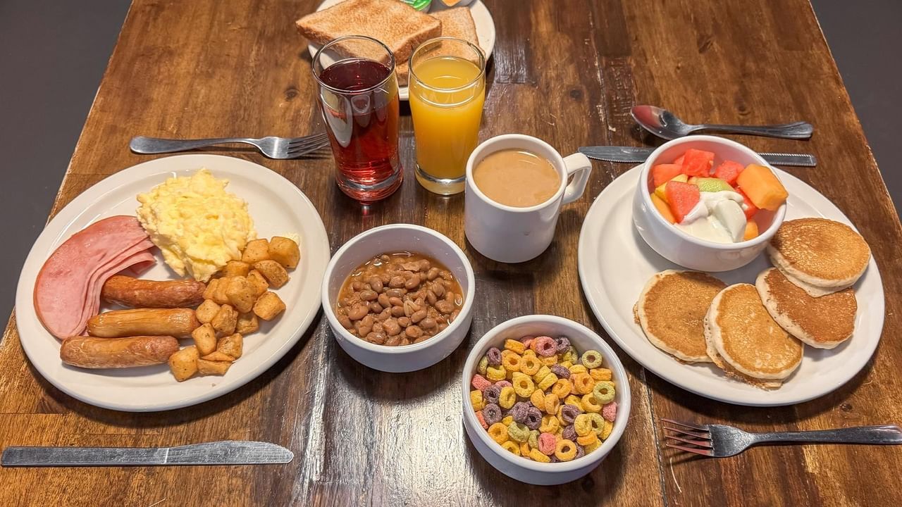 Breakfast spread with pancakes, ham, eggs, beans, cereal, fruit salad, and drinks on a table.