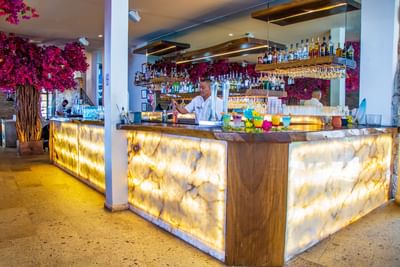 Bar con barra de ónix iluminada, bartender preparando cócteles coloridos y flores fucsia decorativas en Los Arcos Suites