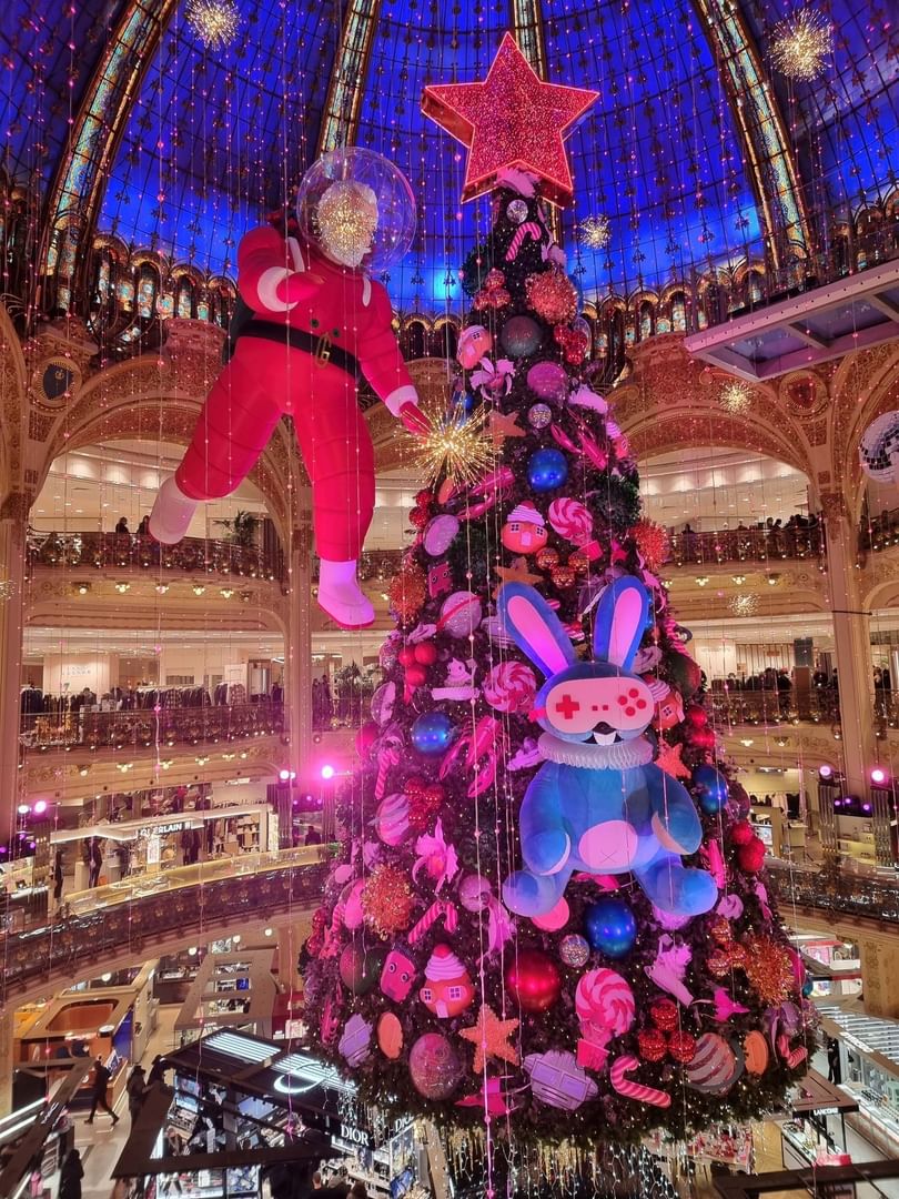 Large Christmas tree in an ornate hall, featuring a hanging red figure and blue rabbit plush at Hotel Westminster Paris