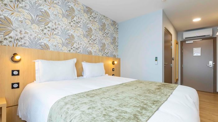 A cozy hotel room with a large bed, floral wallpaper, and a wooden headboard with lighting.