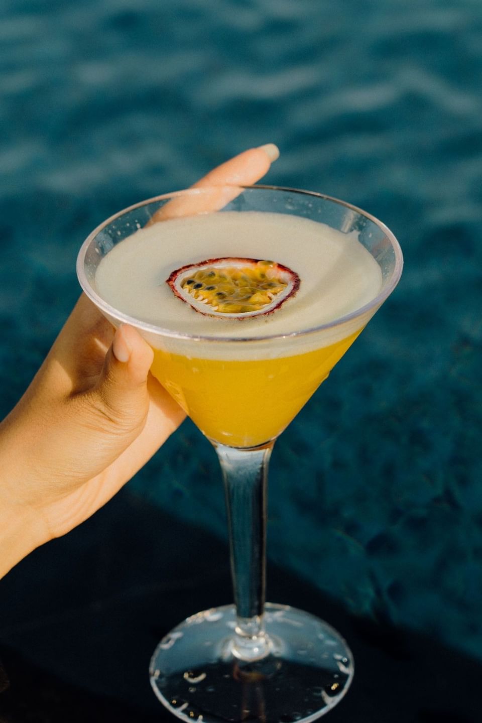 Hand holding a passion fruit martini garnished with fresh fruit against a blue water background at Malibu Sky Lounge & Bar