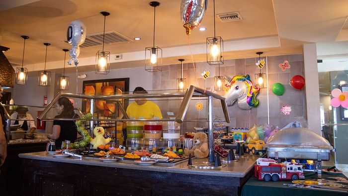 Buffet counter decorated with unicorn balloon and toys under pendant lights in Palmira Cafeteria at Araiza Calafia