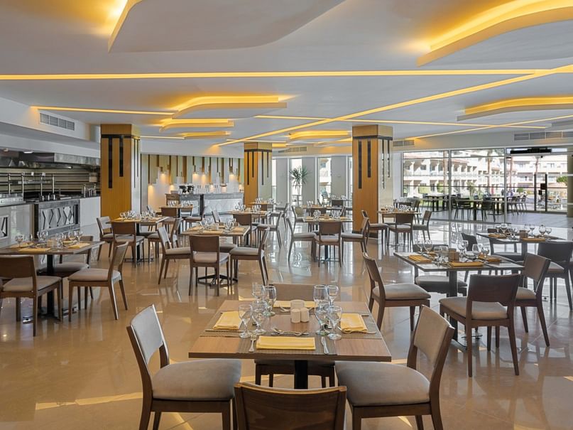 Modern restaurant with dining tables, chairs, and a bar area, illuminated by warm lighting and large windows.