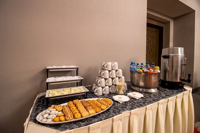 Assorted cookies on a tray by a pyramid of coffee mugs under a tall beverage dispenser on a buffet table at Hotel Mirra
