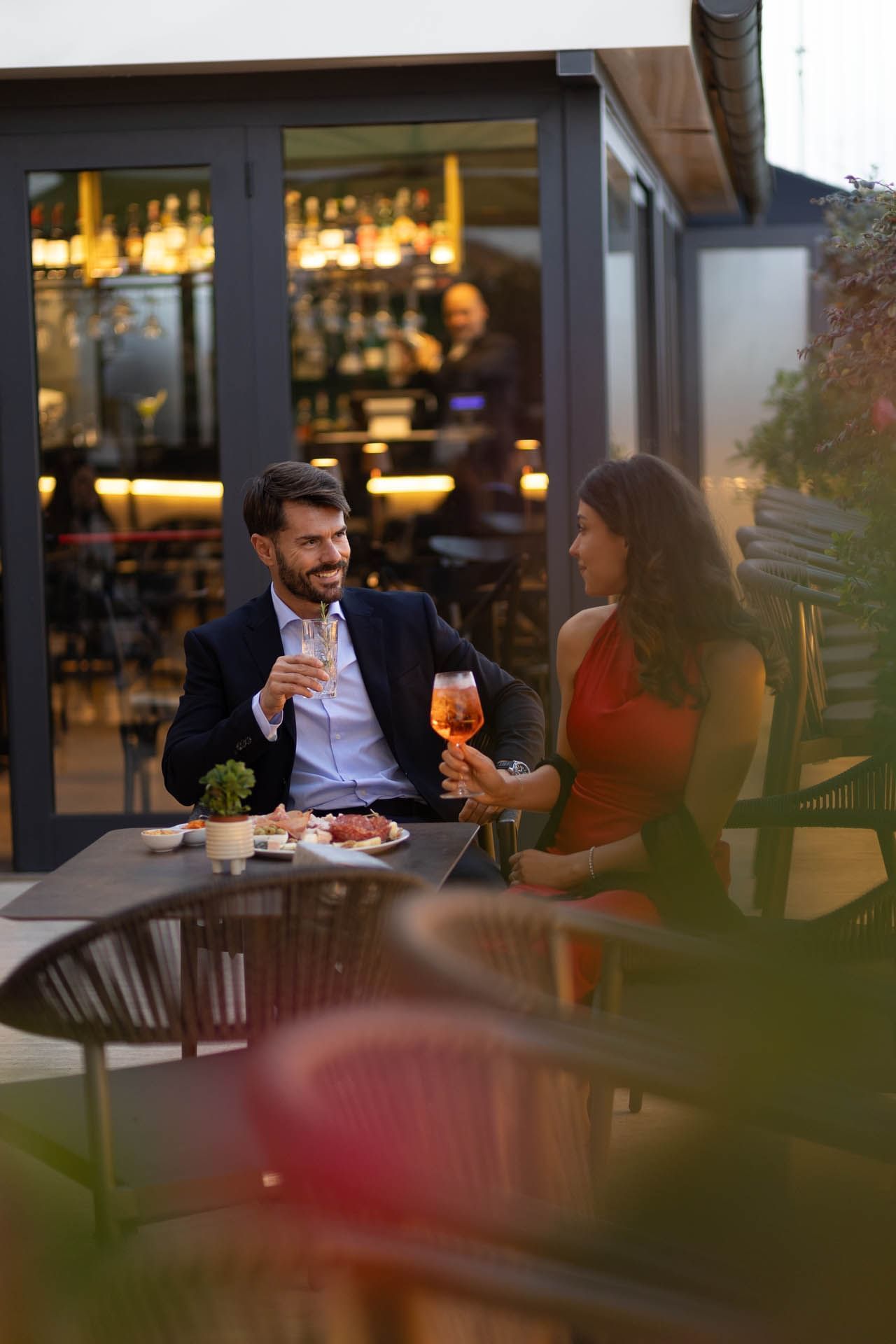 A couple enjoys drinks with food at a table in a stylish outdoor setting, lit by warm evening lights at The Guardian Hotel
