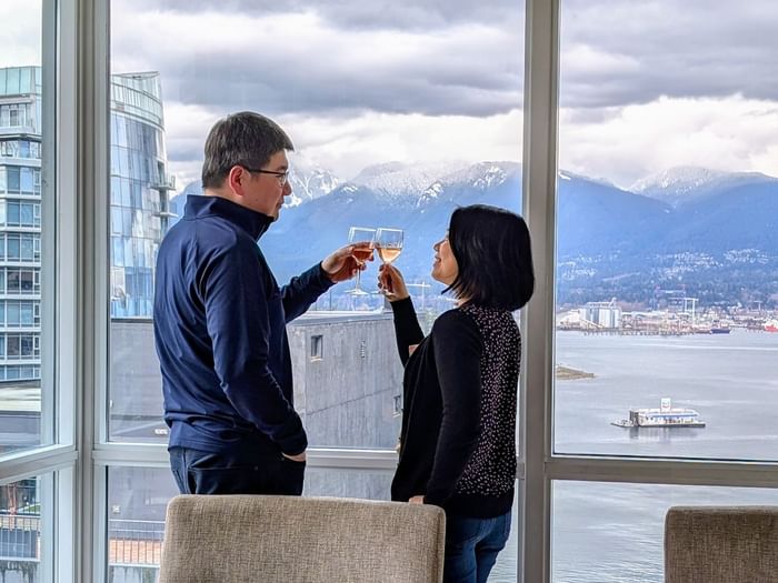 Hotel guests toasting with wine by a window overlooking Vancouver and the ocean.