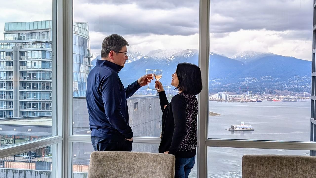 Hotel guests toasting with wine by a window overlooking Vancouver and the ocean.