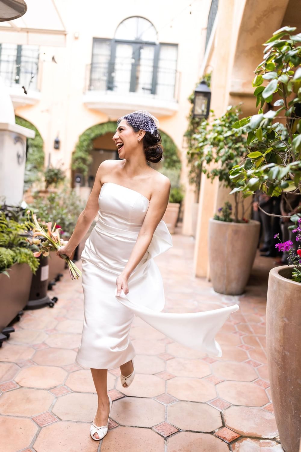 Joyous bride in a white dress and veil, holding a bouquet, walking on a charming pathway at El Prado Hotel