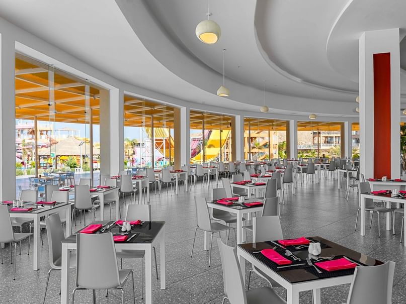 An empty restaurant with many white tables and chairs, pink napkins, and curved ceiling with hanging lights.