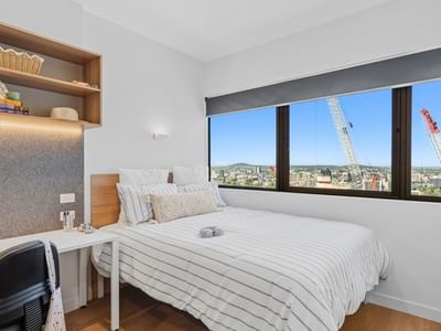 A cozy bedroom with a bed, desk, chair, and large windows at UniLodge Brisbane City.
