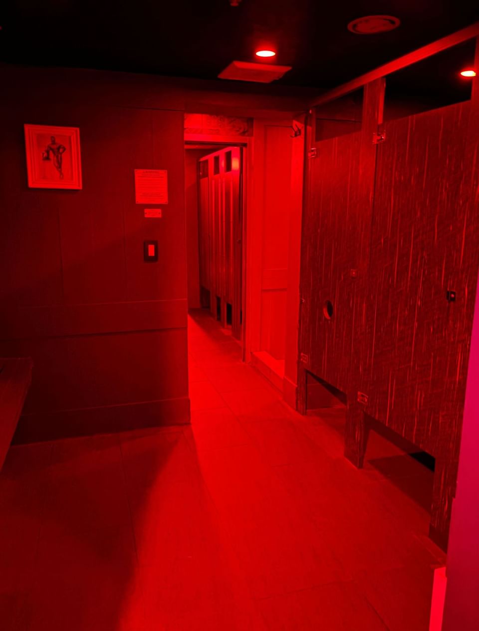 A dimly lit restroom hallway with red lighting, wooden stall doors, and a framed artwork on the wall at The Gaythering
