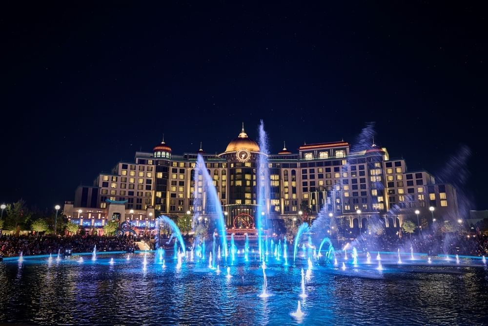 A large hotel at night with glowing windows behind a beautiful blue-lit fountain.