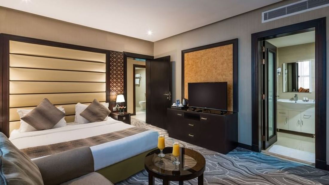 Large bed by a TV stand with two juices on a round table in Executive Suite at Naviti by Warwick Al Khobar