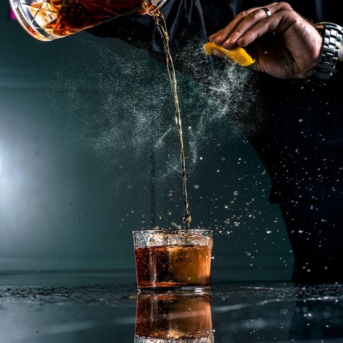 A person pouring whiskey into a glass with a lemon wedge, causing splashes and mist.