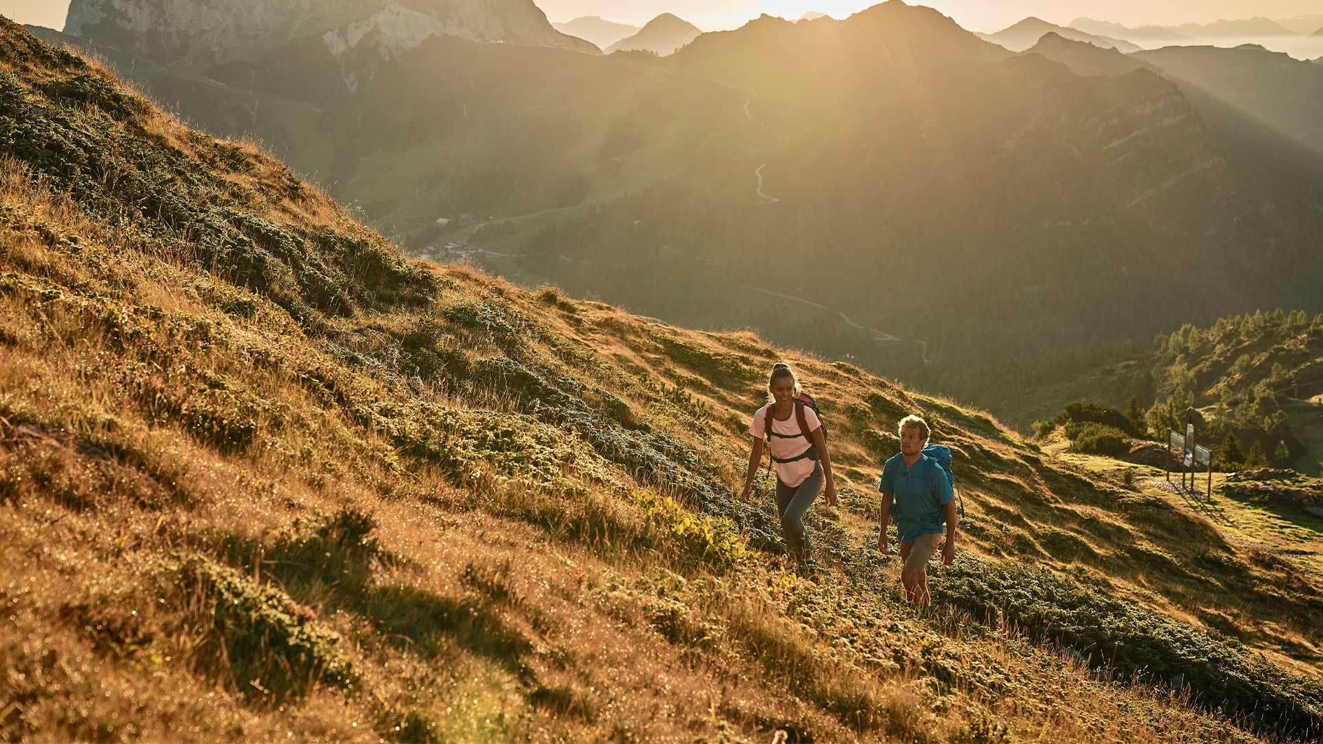 Zwei Wanderer mit Rucksäcken gehen auf einem Bergpfad bei Sonnenaufgang in den Alpen.