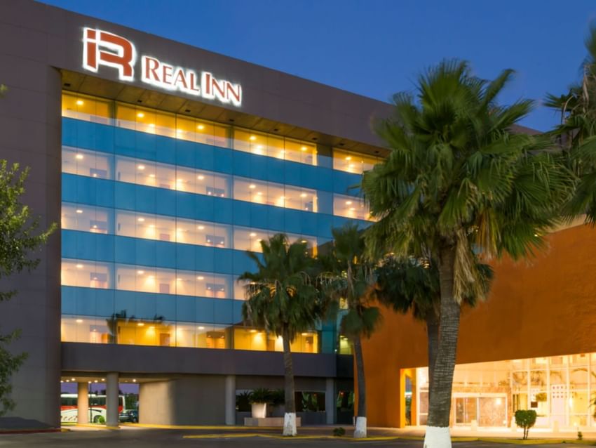 Modern exterior of Real Inn Nuevo Laredo at dusk, showing illuminated windows and tropical palm trees