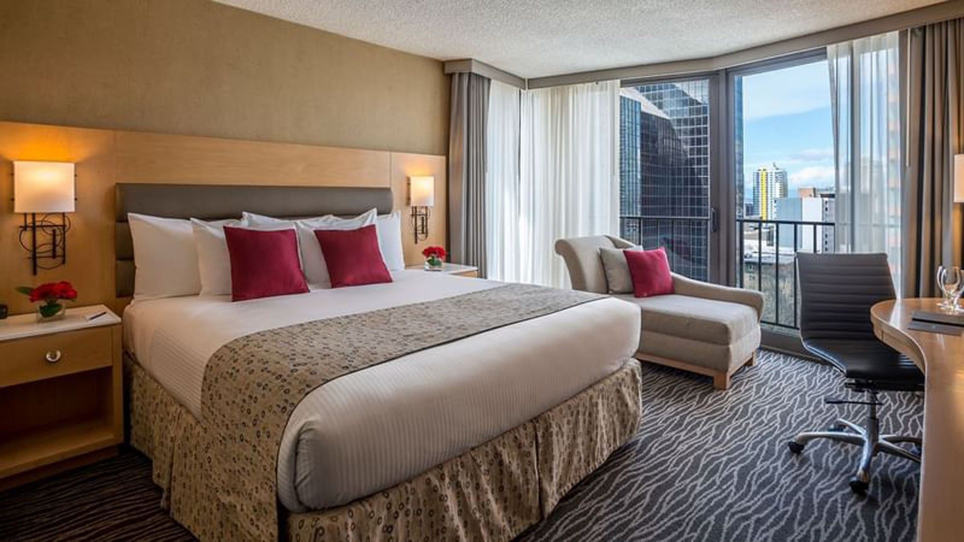 A deluxe hotel room with a king-sized bed, desk, chair, and balcony with a city view.