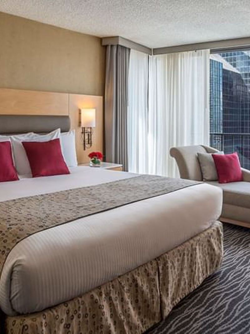 Plush bed by a tan chaise lounge and desk chair surrounding a large glass window in Deluxe room at Warwick Seattle
