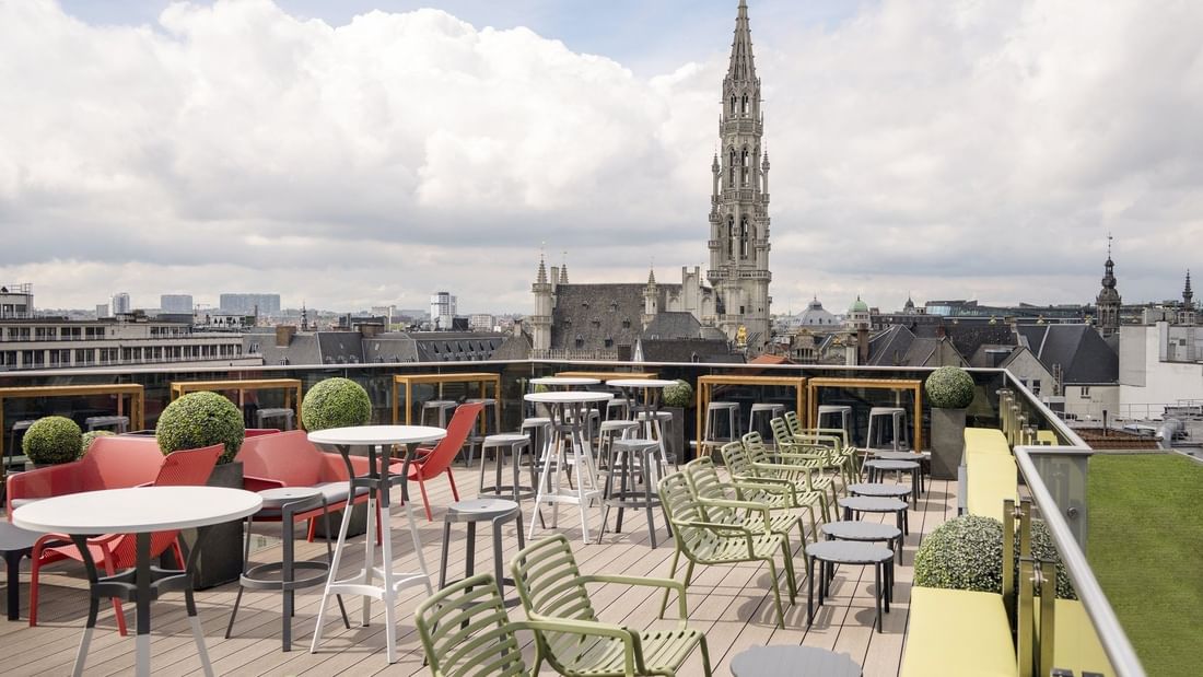 Rooftop terrace with colorful seating, decorative greenery, and panoramic city views featuring a historic Gothic tower.