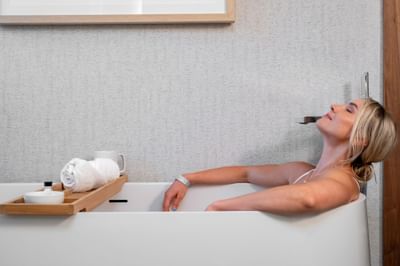 Woman relaxing in a luxurious deep soaking tub, with bath towels and a mug on a tray at Maui Coast Hotel