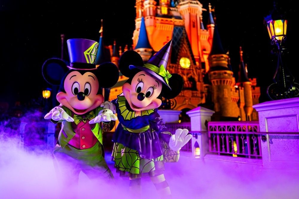 Mickey and Minnie Mouse in their Halloween attire amidst a cloud of fog at night with Cinderella Castle glowing orange in the distance.