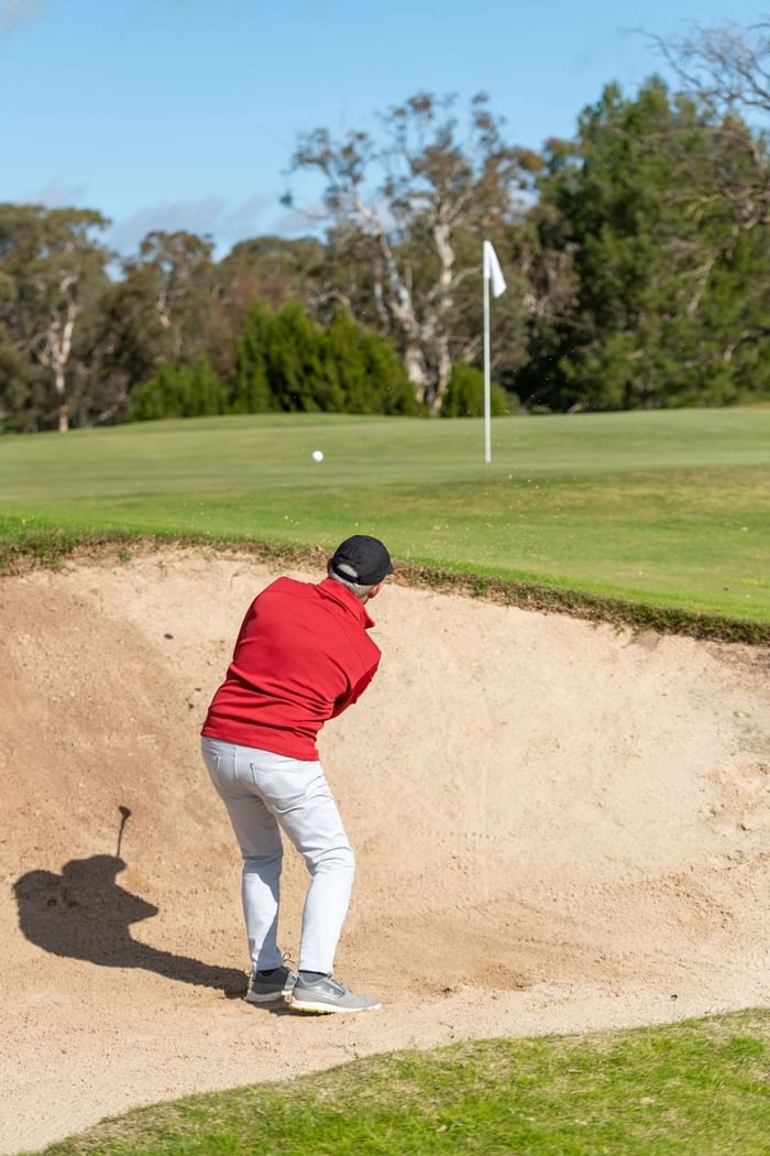 A golfer in a red shirt hitting a ball out of a sand bunker at Novotel Barossa Valley Resort under a clear sky