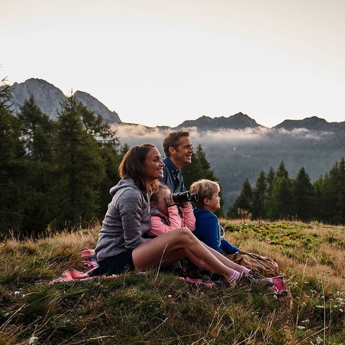 Familie sitzt auf Wiese vor Bergpanorama, Kind mit Fernglas, alle lächeln.